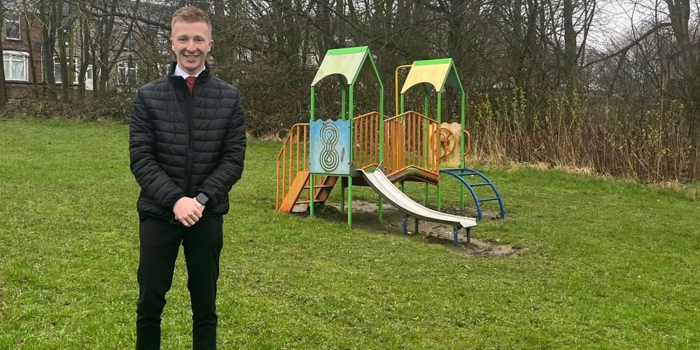 Mayor-Cllr-Curtis-Bihari in a black jacket stands on grass in front of a small, colourful playground structure.