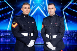 Two men in formal military uniforms pose on a brightly lit stage with a blue star background.