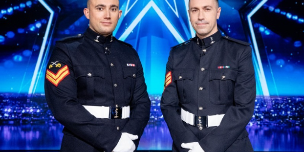 Two men in formal military uniforms pose on a brightly lit stage with a blue star background.
