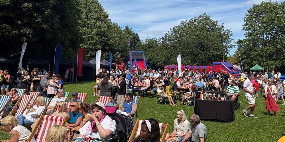 Crowds 2 at Party in the Park 2026 Crowd enjoying a sunny outdoor festival with deckchairs, food stalls, and inflatable slides.