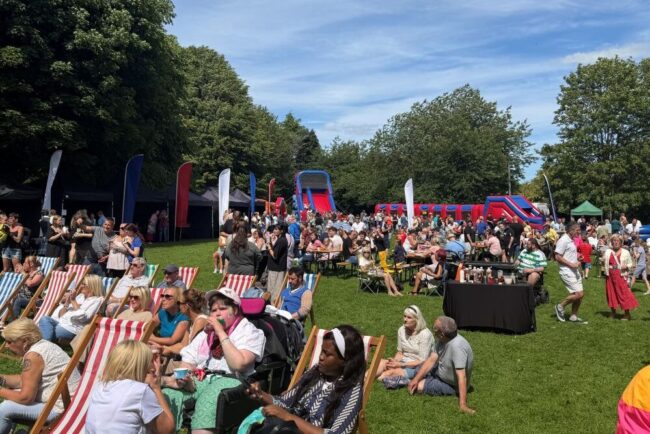 Crowd enjoying a sunny outdoor festival with deckchairs, food stalls, and inflatable slides.