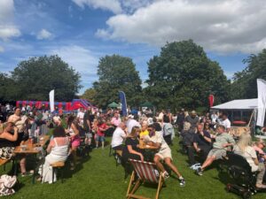 A large crowd gathers at an outdoor festival with marquees, tables, and inflatables under sunny skies.
