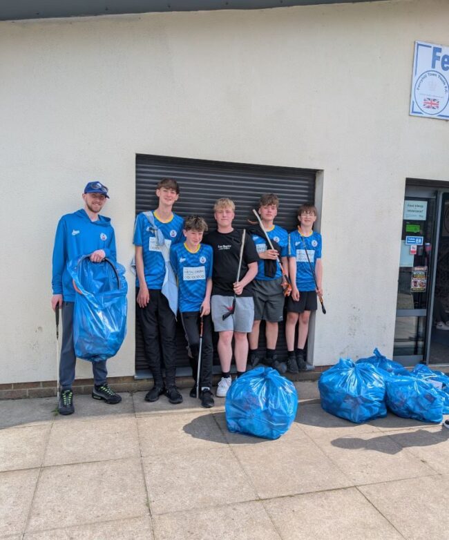 Six people in blue shirts stand by blue bin bags outside a building after tidying up.