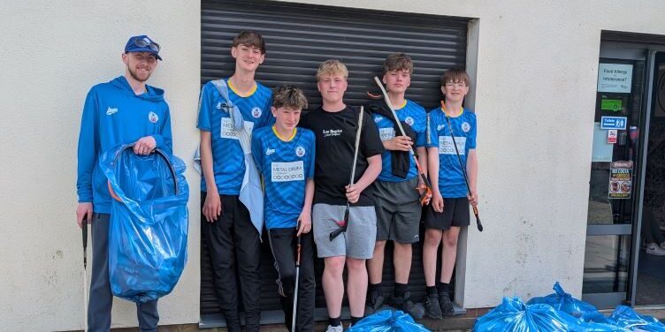 Six people in blue shirts stand by blue bin bags outside a building after tidying up.