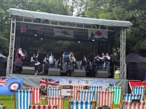 A pipe band performs on an outdoor stage with empty striped deckchairs in front.