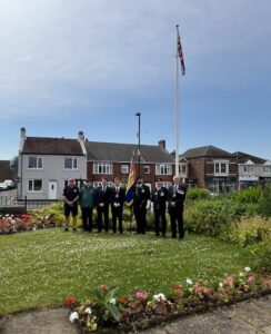 Seven people stand in a garden with flags, flowers, and houses in the background.
