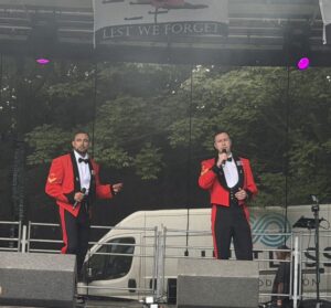 Two men in red military uniforms singing on an outdoor stage, with a Lest We Forget banner above.