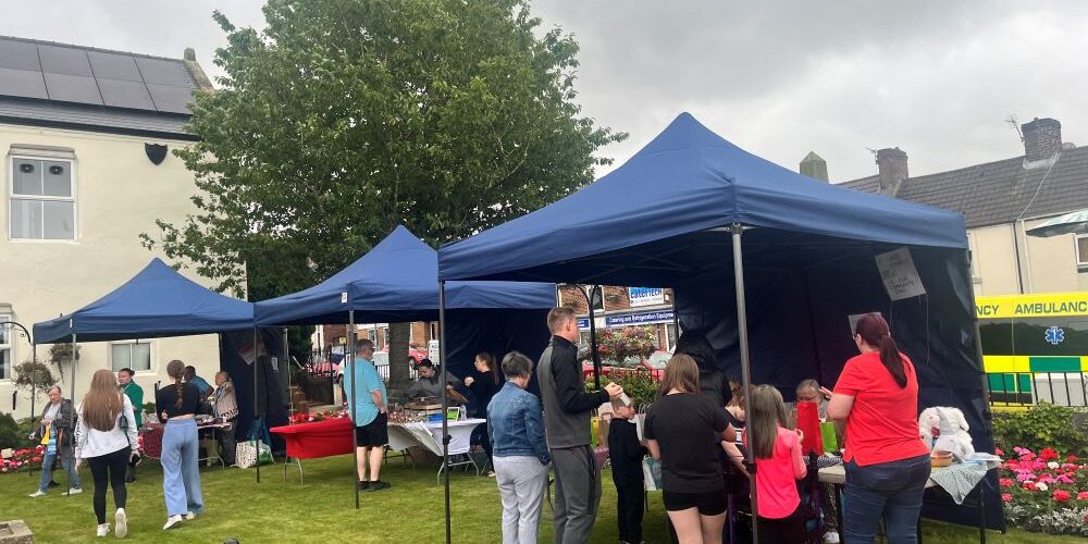 People gather under blue marquees at an outdoor fete with flowers and an ambulance in the background.