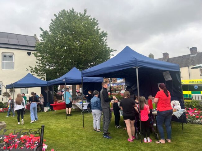 People gather under blue marquees at an outdoor fete with flowers and an ambulance in the background.