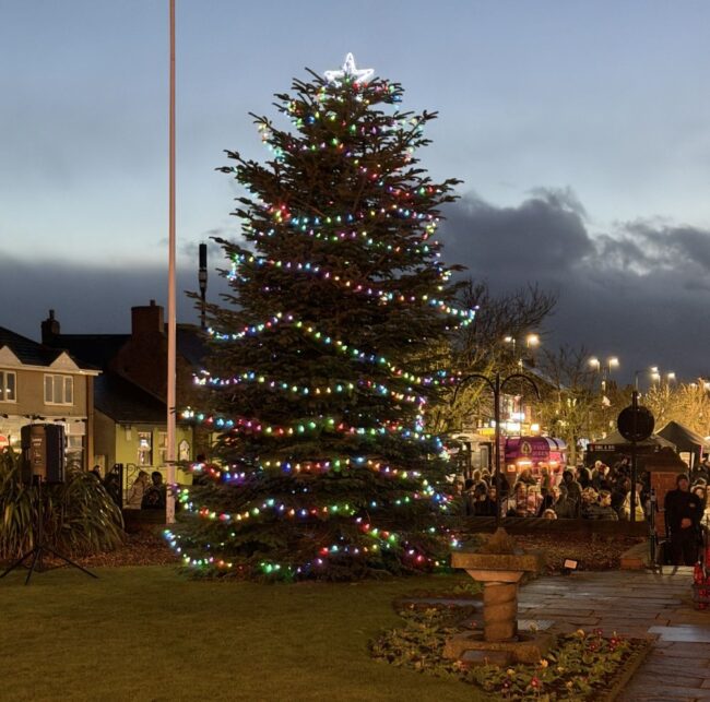 Large Christmas tree with colourful lights stands in a town square at dusk.