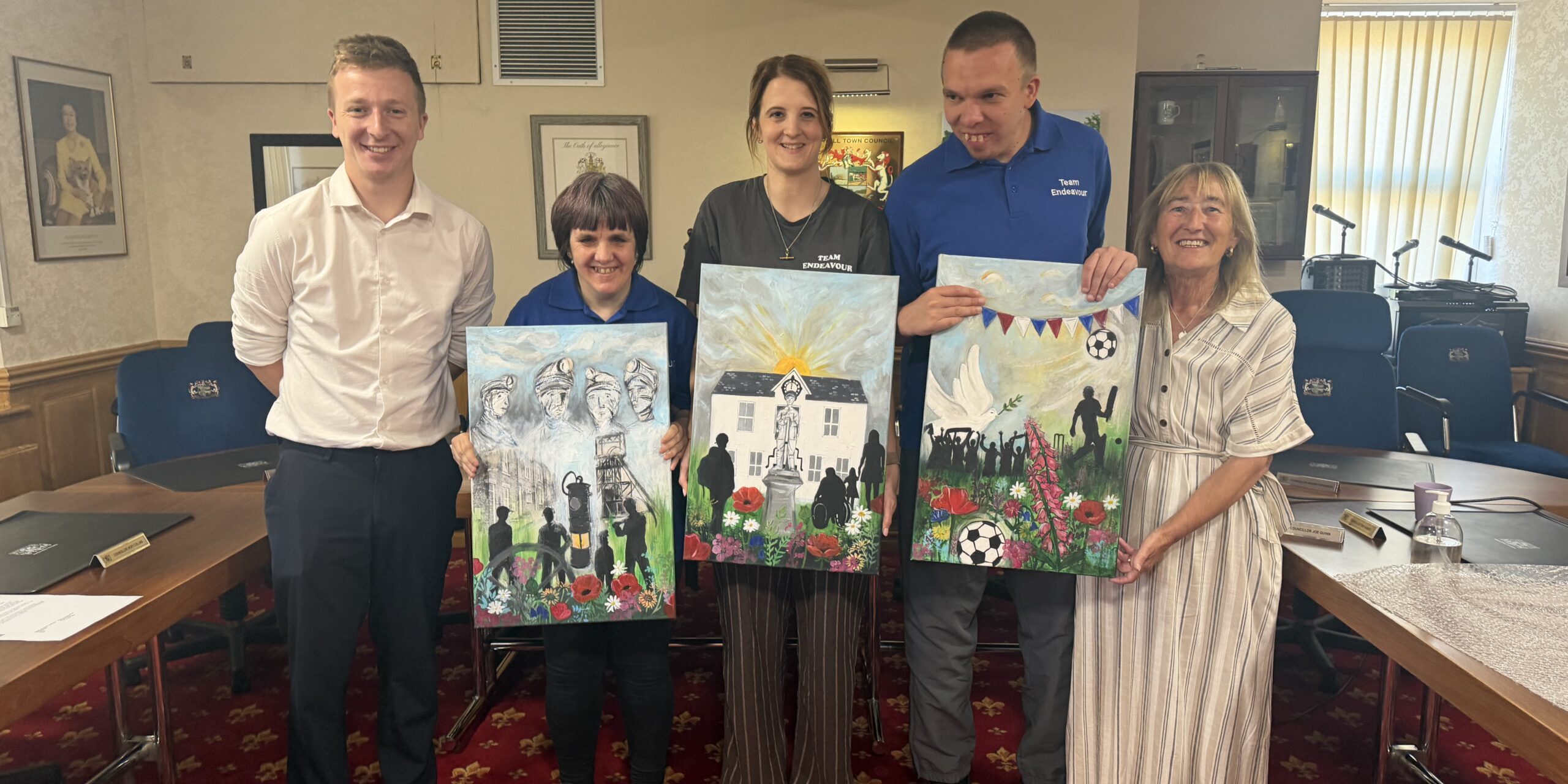 Five people indoors smiling and holding up colourful paintings in a formal meeting room.
