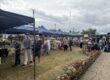 People browsing stalls at an outdoor market, with flowers and marquees under a cloudy sky.