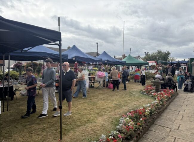 People browsing stalls at an outdoor market, with flowers and marquees under a cloudy sky.