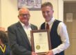 Two men pose holding a framed certificate, smiling, with people seated in the background.