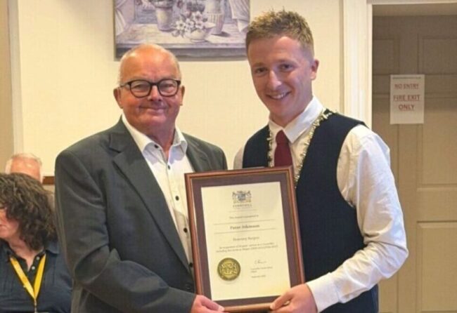 Two men pose holding a framed certificate, smiling, with people seated in the background.