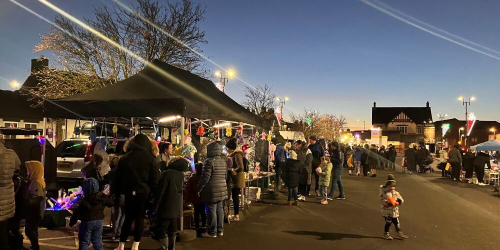 People gather at outdoor market stalls at dusk, with lights and traders under marquees.