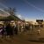People gather at outdoor market stalls at dusk, with lights and traders under marquees.