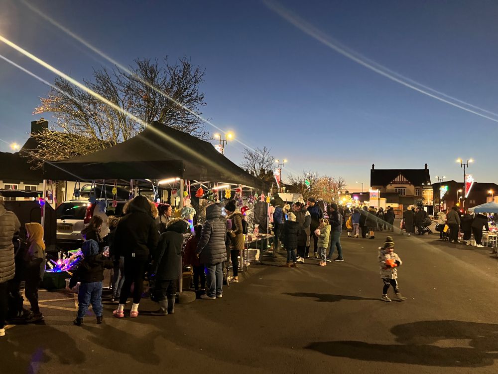 People gather at outdoor market stalls at dusk, with lights and traders under marquees.