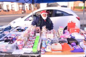 A vendor in a Santa hat sits behind a colourful holiday market stall on a sunny street.